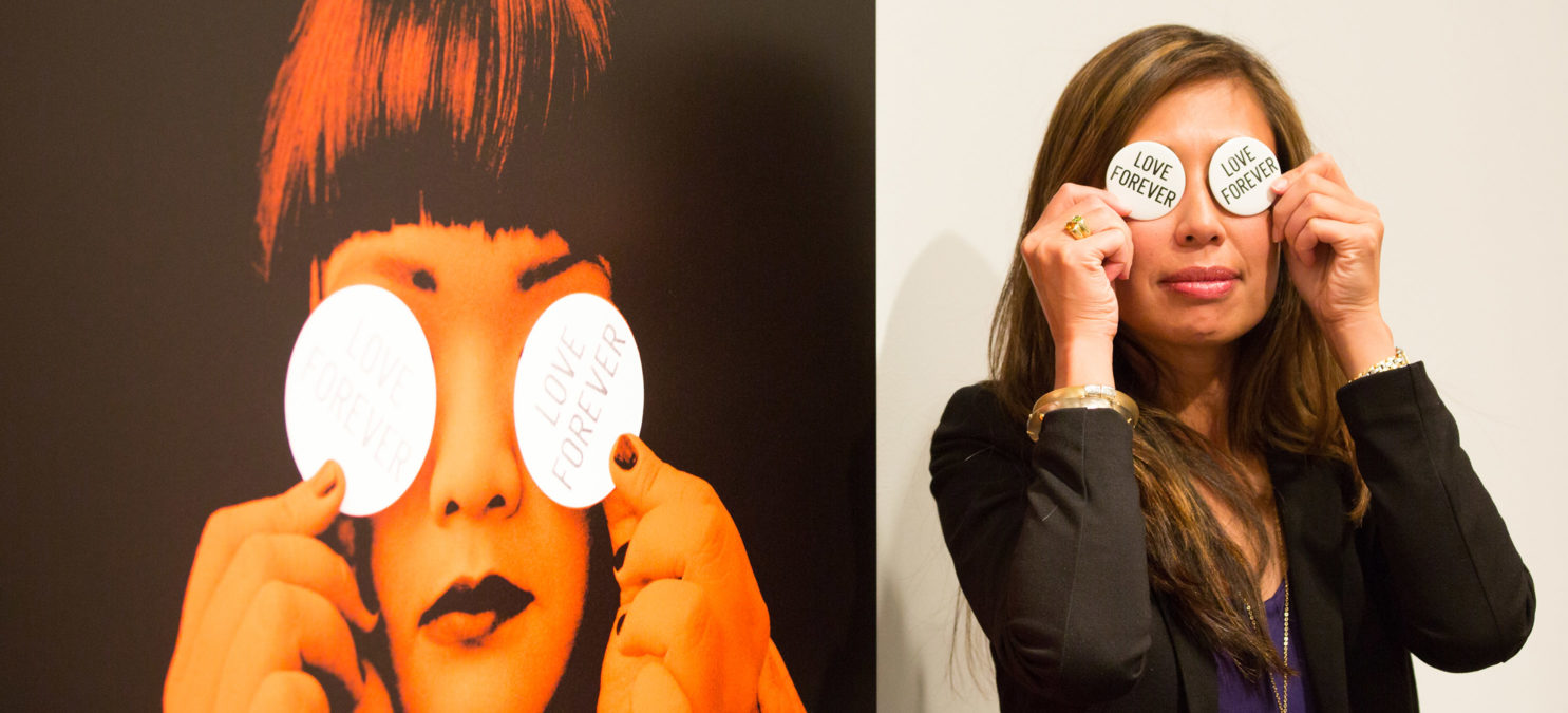 A woman holds two paper circles with "Love Forever" printed on them over her eyes, mimicking the artwork beside her